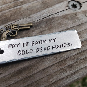 May include: A silver metal keychain with a small gun charm and the text "Pry it from my cold dead hands."