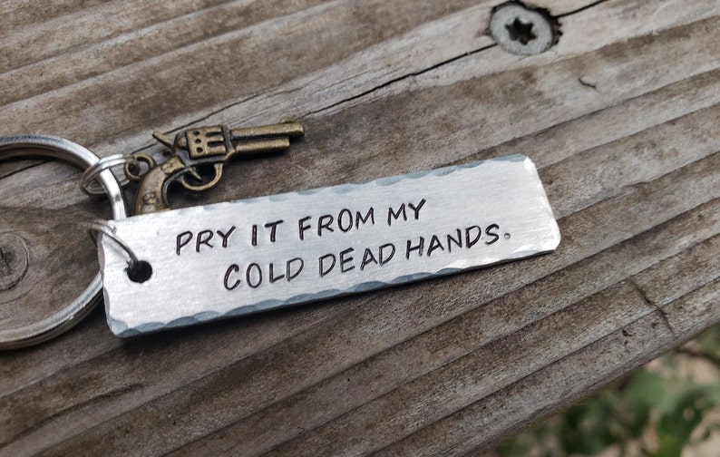 May include: A silver metal keychain with a small gun charm and the text "Pry it from my cold dead hands."