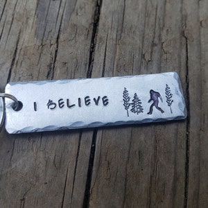 Op de afbeelding: Een zilveren sleutelhanger met de woorden "I Believe" en een silhouet van Bigfoot die door een bos loopt.