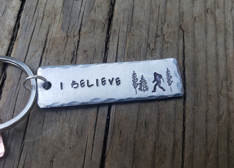 Op de afbeelding: Een zilveren sleutelhanger met de woorden "I Believe" en een silhouet van Bigfoot die door een bos loopt.