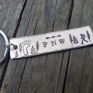 Op de afbeelding: Een zilveren metalen sleutelhanger met een rechthoekig label gegraveerd met de woorden "PNW" en afbeeldingen van een voetafdruk, bomen en een Bigfoot.
