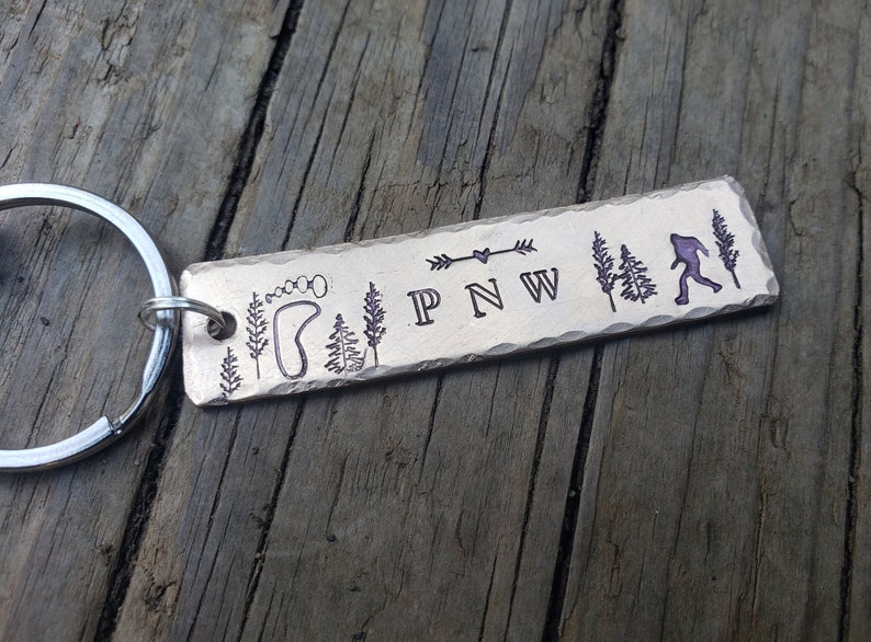 Op de afbeelding: Een zilveren metalen sleutelhanger met een rechthoekig label gegraveerd met de woorden "PNW" en afbeeldingen van een voetafdruk, bomen en een Bigfoot.