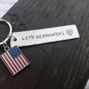 May include: A silver keychain with a small American flag charm and a metal tag that reads "LET'S GO BRANDON!" with a small American flag heart.
