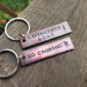 Op de afbeelding: Twee metalen sleutelhangers met gegraveerde tekst. De eerste sleutelhanger zegt "Livingston Road" met een klein boomontwerp. De tweede sleutelhanger zegt "Go Camping!" met een stick figure die wandelt.