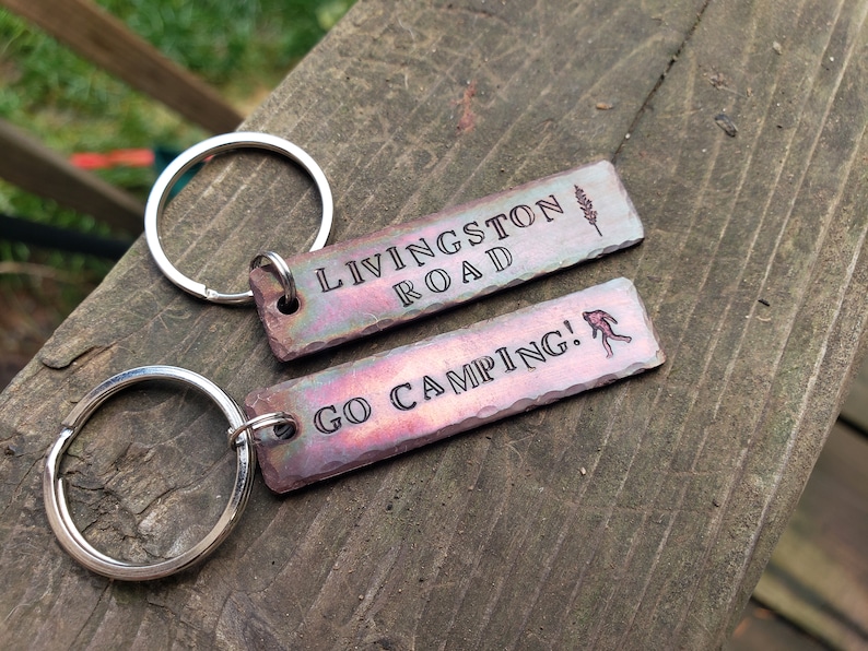 Op de afbeelding: Twee metalen sleutelhangers met gegraveerde tekst. De eerste sleutelhanger zegt "Livingston Road" met een klein boomontwerp. De tweede sleutelhanger zegt "Go Camping!" met een stick figure die wandelt.