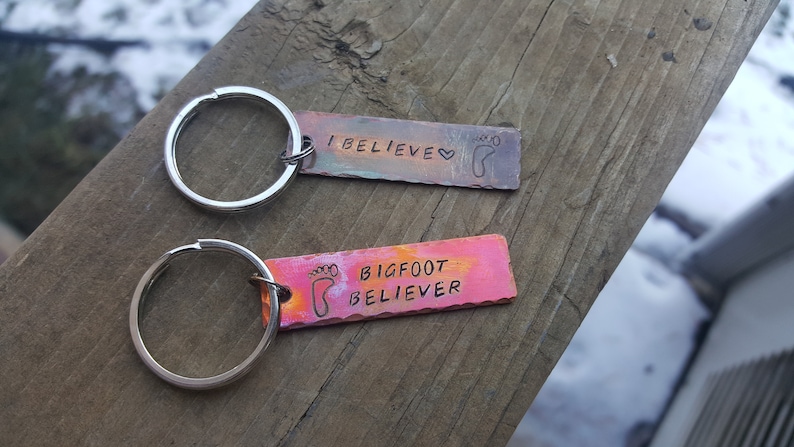 Op de afbeelding: Twee metalen sleutelhangers met een zilveren sleutelring. De bovenste sleutelhanger heeft een kleurrijk rechthoekig label met de tekst "I BELIEVE" en een voetafdruk. De onderste sleutelhanger heeft een kleurrijk rechthoekig label met de tekst "BIGFOOT BELIEVER" en een voetafdruk.