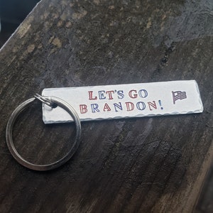 May include: A silver keychain with a white rectangular tag that says "Let's Go Brandon!" in red and blue lettering with a small American flag graphic.