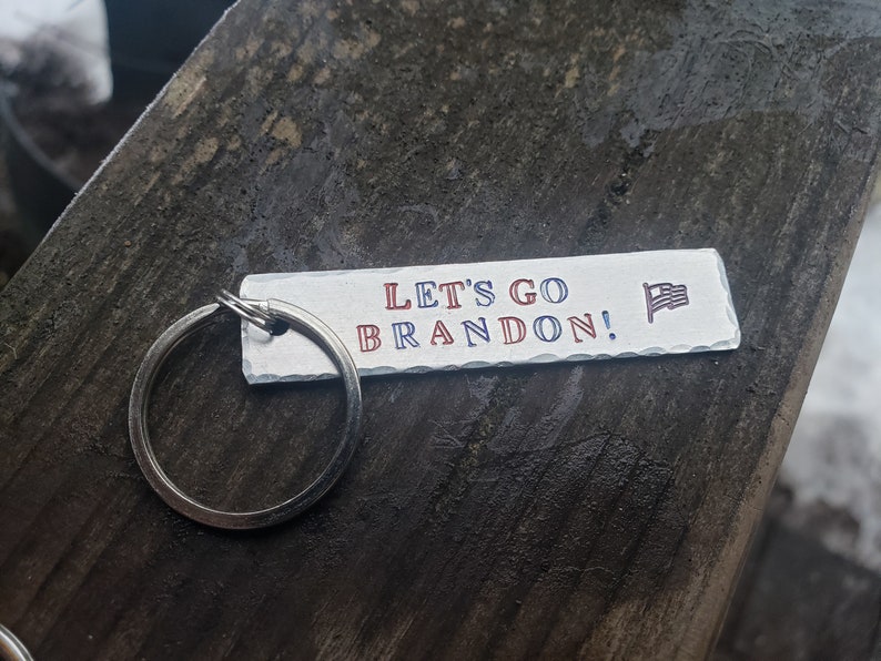 May include: A silver keychain with a white rectangular tag that says "Let's Go Brandon!" in red and blue lettering with a small American flag graphic.