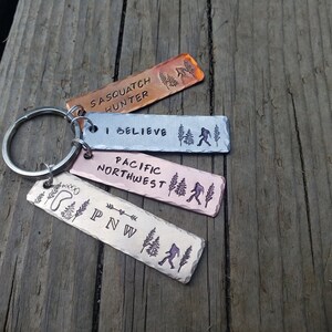 Op de afbeelding: Een set van vier metalen sleutelhangers met verschillende ontwerpen. De sleutelhangers zijn allemaal rechthoekig en hebben een zilveren ring. De ontwerpen omvatten "Sasquatch Hunter", "I Believe", "Pacific Northwest" en "PNW".