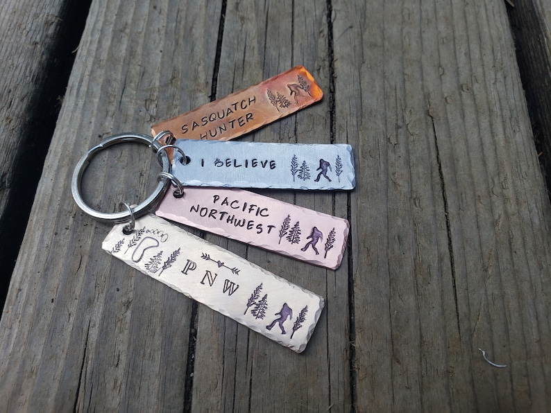 Op de afbeelding: Een set van vier metalen sleutelhangers met verschillende ontwerpen. De sleutelhangers zijn allemaal rechthoekig en hebben een zilveren ring. De ontwerpen omvatten "Sasquatch Hunter", "I Believe", "Pacific Northwest" en "PNW".