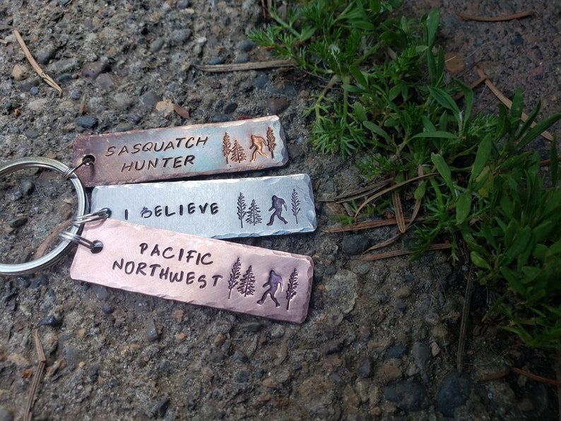 Op de afbeelding: Drie metalen sleutelhangers met verschillende ontwerpen. De bovenste sleutelhanger is koperkleurig en heeft de tekst "Sasquatch Hunter" met een silhouet van Bigfoot en dennenbomen. De middelste sleutelhanger is zilverkleurig en heeft de tekst "Ik geloof" met een silhouet van Bigfoot en dennenbomen. De onderste sleutelhanger is koperkleurig en heeft de tekst "Pacific Northwest" met een silhouet van Bigfoot en dennenbomen.