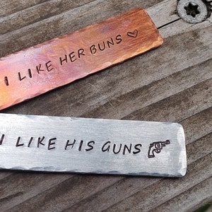 May include: Two metal keychains with engraved text. The top keychain is copper and reads "I like her buns" with a heart symbol. The bottom keychain is silver and reads "I like his guns" with a small gun symbol.