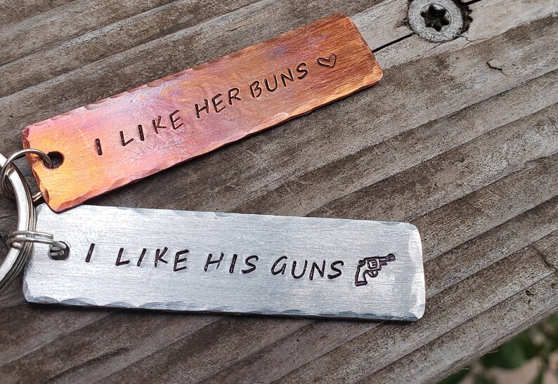 May include: Two metal keychains with engraved text. The top keychain is copper and reads "I like her buns" with a heart symbol. The bottom keychain is silver and reads "I like his guns" with a small gun symbol.