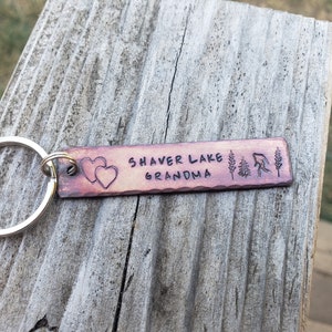 Op de afbeelding: Een koperen sleutelhanger met de tekst "Shaver Lake Grandma" en twee harten. De sleutelhanger heeft ook een kleine boom en een vis.