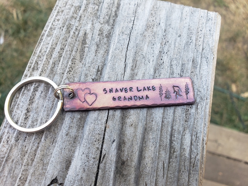 Op de afbeelding: Een koperen sleutelhanger met de tekst "Shaver Lake Grandma" en twee harten. De sleutelhanger heeft ook een kleine boom en een vis.