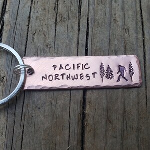Op de afbeelding: Een koperen sleutelhanger met de woorden "PACIFIC NORTHWEST" erop gegraveerd. De sleutelhanger heeft ook een silhouet van een persoon die loopt en twee dennenbomen.