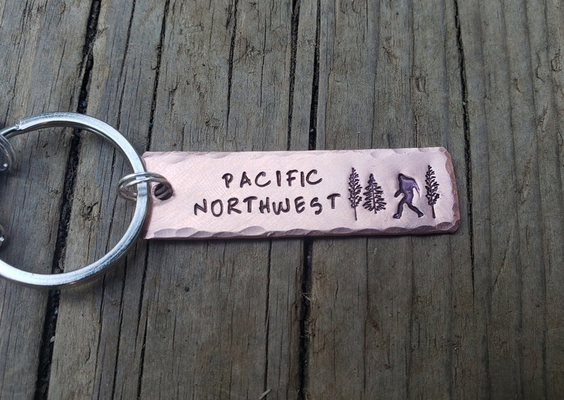 Op de afbeelding: Een koperen sleutelhanger met de woorden "PACIFIC NORTHWEST" erop gegraveerd. De sleutelhanger heeft ook een silhouet van een persoon die loopt en twee dennenbomen.