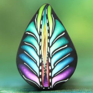 Op de afbeelding: Een traanvormige kralen van polymer clay met een kleurrijk, abstract ontwerp. De kraal heeft een centrale, verticale lijn met een reeks gebogen lijnen die naar buiten stralen, waardoor een veervormig patroon ontstaat. De kleuren zijn levendig en omvatten tinten groen, blauw, paars en wit.
