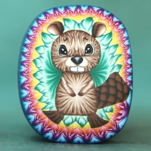 May include: Oval-shaped artwork featuring a cartoon beaver with brown fur, large eyes, and a checkered tail. The beaver is centered within a vibrant, rainbow-colored floral design, set against a teal background. The artwork is likely a decorative item.