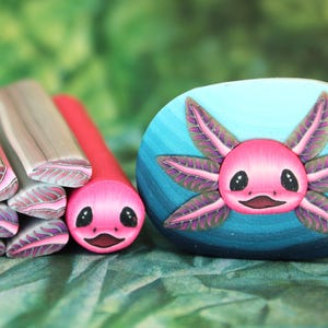 May include: A collection of polymer clay axolotl creations. The image features a pink axolotl face on a blue stone, along with several axolotl-themed clay canes in pink, gray, and red. The axolotls have black eyes and smiling mouths.