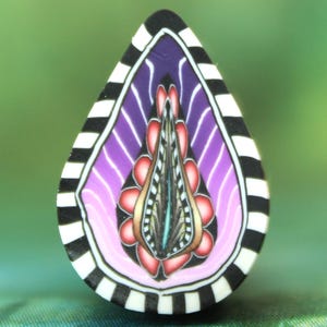 Op de afbeelding: Een traanvormige hanger met een zwart-wit gestreepte rand. Het midden van de hanger heeft een paarse en roze gradiënt met een gedetailleerd bloemmotief in tinten rood, roze en zwart.