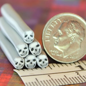 Op de afbeelding: Zes grijze, schedelvormige kralen met zwarte details zijn naast een munt en een liniaal gestapeld. De kralen zijn cilindrisch met een schedelontwerp aan het uiteinde.