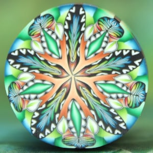 May include: A colorful, round polymer clay creation with a symmetrical floral design. The artwork features a central orange starburst pattern, surrounded by green, blue, and white leaf-like shapes. The edges are a mix of blue and green.
