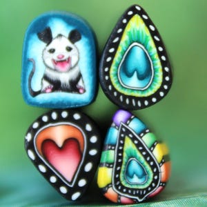 Op de afbeelding: Vier kleurrijke, handgemaakte polymeerklei hangers. Eén heeft een opossum, een andere een hart en twee hebben druppelvormige ontwerpen met ingewikkelde patronen. De hangers zijn geplaatst tegen een groene achtergrond.