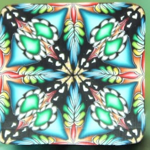 May include: Square, decorative object with a symmetrical, floral-like design. The pattern features teal, green, orange, and yellow elements against a black background. White heart-shaped accents add detail to the design. The object has rounded corners.
