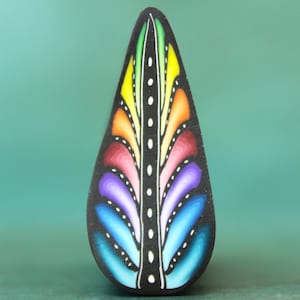 May include: A teardrop-shaped, black pendant with a colorful, stylized feather design. The feather features a spectrum of colors, including blue, purple, red, orange, and yellow, with white accents. The pendant is likely a piece of jewelry.