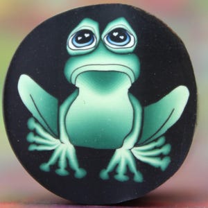 May include: A green frog with big, blue eyes and a sad expression is painted on a black round surface. The frog has a white heart shape in each eye.