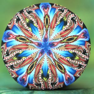 May include: A round, decorative object with a vibrant, symmetrical design. The pattern features a central blue starburst, surrounded by red, brown, and black petal-like shapes against a blue and black background. The object is set against a blurred green backdrop.
