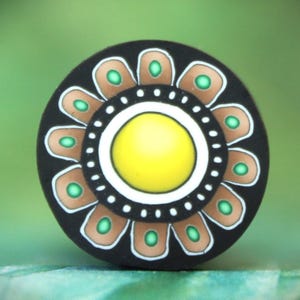 May include: A round, black decorative object with a floral design. The center is yellow, surrounded by a white ring, a ring of brown petals with green accents, and a black outer edge. The background is a soft green.