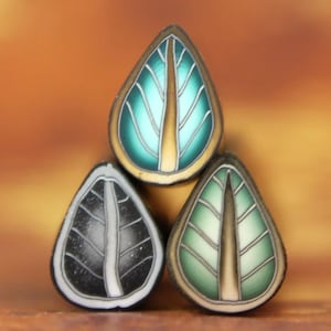 Op de afbeelding: Drie keramische hangers in de vorm van een blad met een gouden en blauw ontwerp. De hangers zijn op elkaar gestapeld.