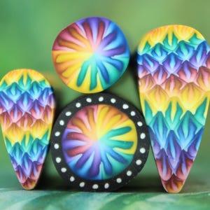 May include: A collection of colorful polymer clay beads in various shapes. The beads feature a rainbow spiral design and a textured pattern resembling scales. One bead has a black border with white dots.