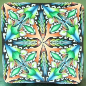 Op de afbeelding: Een vierkant decoratief object met een kleurrijk, symmetrisch bloemmotief. Het ontwerp bevat oranje, groene, blauwe en witte elementen op een zwarte achtergrond. Het object heeft afgeronde randen en een glanzende afwerking.