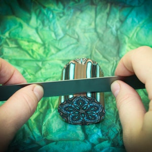 May include: A person's hands are using a metal file to smooth the edges of a colorful, cylindrical polymer clay pendant. The pendant has a geometric pattern with white, brown, blue, and black colors.