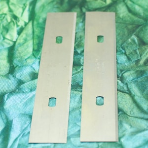 May include: Two silver-colored razor blades with three holes each. The blades are on a green and blue background.