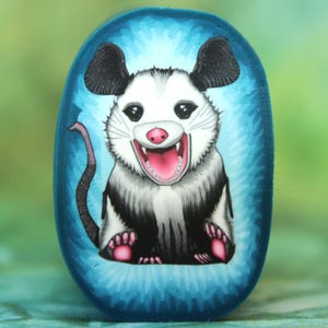 Op de afbeelding: Een handbeschilderde, ovaalvormige steen met een cartoonbuidelrat. De buidelrat heeft zwart-witte vacht, roze poten en een roze neus. Het is geplaatst tegen een blauwe achtergrond met stralende lijnen, wat de indruk van een schijnwerper wekt.