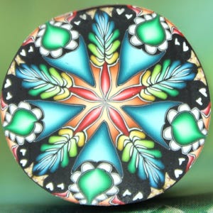 Op de afbeelding: Een ronde, kleurrijke polymeerklei hanger met een bloemmotief. De hanger heeft een zwarte achtergrond met een centraal sterpatroon in rood, oranje, geel en turquoise. Groene, witte en blauwe bladvormen en bloemblaadjes completeren het ontwerp.