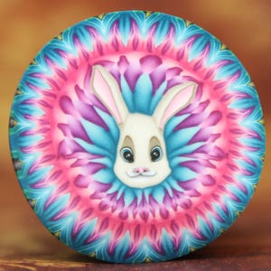 Op de afbeelding: Een wit konijn met roze oren en blauwe ogen is omringd door een kleurrijk mandala-patroon in tinten roze, blauw en groen.