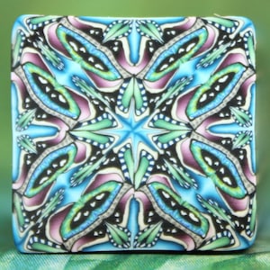 May include: A square tile with a kaleidoscope pattern in shades of blue, green, pink, and black. The pattern is symmetrical and features a star in the center.
