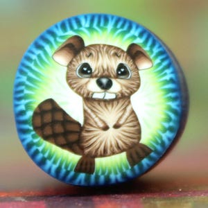 May include: A round, colorful button featuring a cartoon beaver. The beaver is brown with a white belly, a black nose, and large, expressive eyes. The background is a tie-dye pattern of blue, green, and yellow.