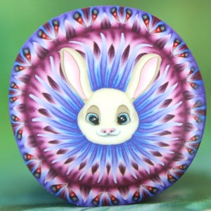 Op de afbeelding: Een rond decoratief item met een wit konijnengezicht in het midden, blauwe ogen en roze accenten. Het konijn is omgeven door een mandala-patroon in paarse, blauwe en rode tinten. Het item heeft een glanzende afwerking.