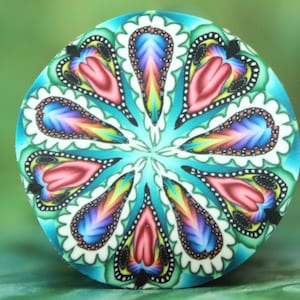 May include: A round, colorful polymer clay design with a floral pattern. The design features teal, pink, purple, and yellow teardrop shapes with white scalloped edges. The center is a vibrant teal color.
