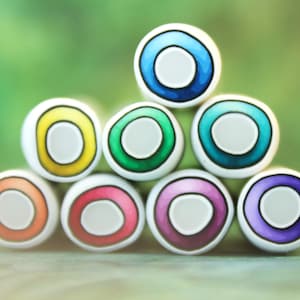 May include: A stack of seven cylindrical beads arranged in a pyramid formation. Each bead features a white outer ring, a vibrant colored inner ring in shades of blue, yellow, green, teal, orange, pink, and purple, and a solid light gray center.