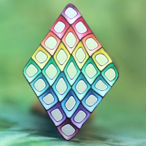 May include: A diamond-shaped brooch featuring a repeating grid pattern of small, light-colored lozenges outlined in a vibrant rainbow gradient. The colors transition smoothly from red and orange at the top to yellow, green, blue, and purple at the base.