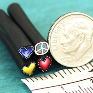 Op de afbeelding: Vier zwarte polymer clay nagelkunststokjes met verschillende ontwerpen. De ontwerpen omvatten een ​​blauw hart, een vredesteken, een geel hart en een rood hart. De stokjes liggen naast een Amerikaanse dime voor schaal.