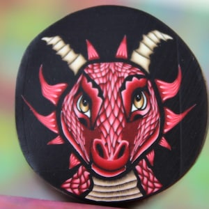 Op de afbeelding: Een rode draak met gele ogen en witte hoorns is geschilderd op een zwarte ronde achtergrond.