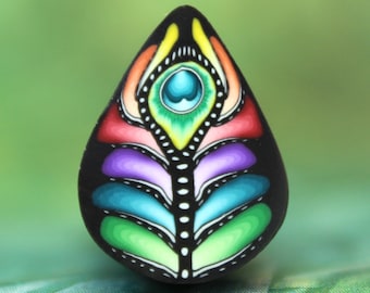 May include: A teardrop-shaped pendant with a black base, featuring a colorful peacock feather design. The design includes shades of yellow, orange, red, purple, blue, and green, with white dotted accents. The pendant has a glossy finish.